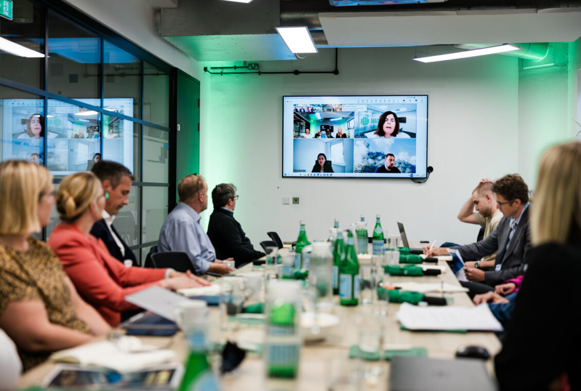 Group working around a large meeting table with people also joining remotely on screen